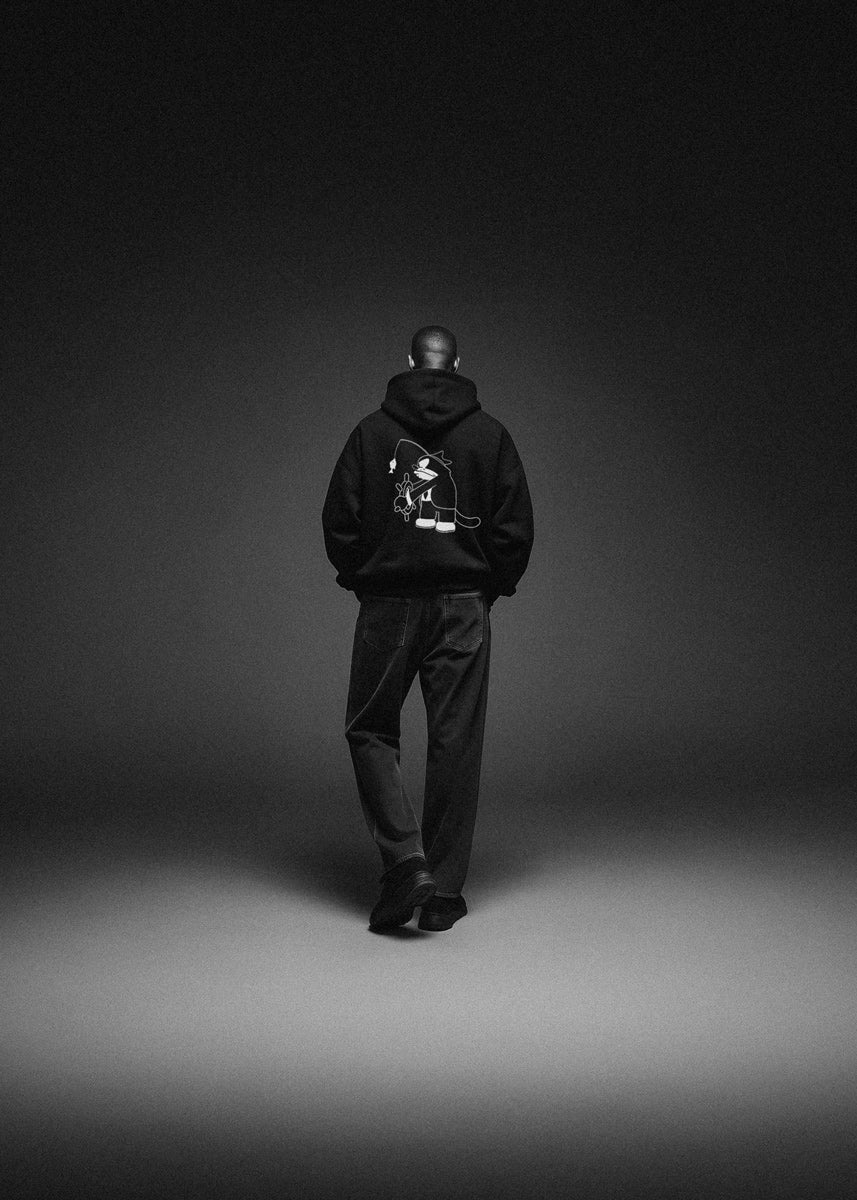 Person wearing a black hoodie with a logo on the back, standing against a dark background